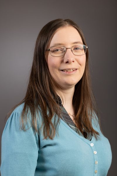 A headshot of Katherine Butler, who has long, straight brown hair, blue eye, wear glasses. She is wearing a teal cardigan over a teal and grey striped shirt.